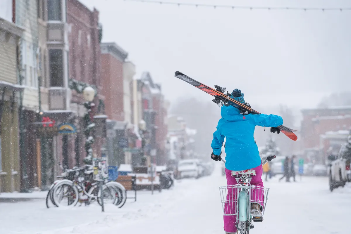 A lively winter street scene with a person on a bike carrying skis, snow-covered streets, and colorful buildings. The image combines biking, skiing, and exploring the town, perfectly illustrating the multi-season, multi-activity access described in the content.