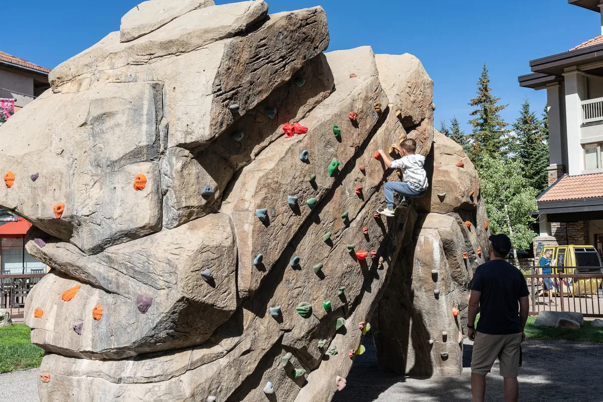 An exterior shot of a child climbing a rock under the watchful eye of an adult. The scene evokes a sense of adventurous play and family interaction, with the outdoors prominently featured, signaling direct access to trails and activity spaces.