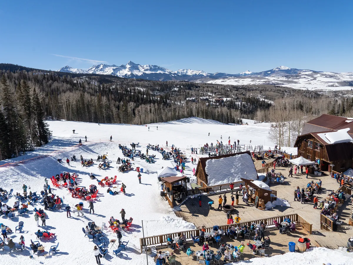 This image captures people enjoying outdoor seating at what appears to be a ski resort base, surrounded by wooden structures and with the stunning mountain backdrop. Groups are relaxing, sharing meals and drinks, and basking in the lively, social energy of the mountainside setting.