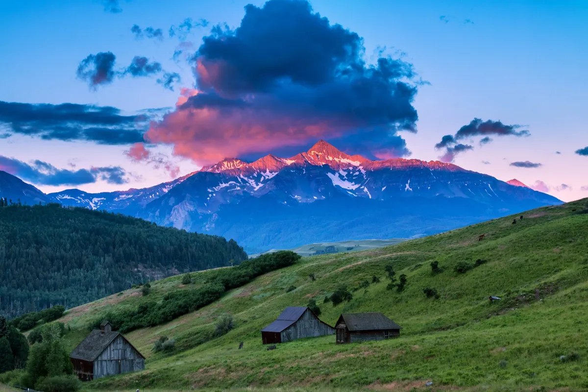 A breathtaking view of snow-capped mountains with rustic cabins nestled below and a vibrant sky overhead. The tranquil landscape creates a sense of peaceful seclusion, ideal for romantic getaways and quiet reflection. The setting suggests opportunities for hiking, photography, and enjoying the serene beauty of nature.