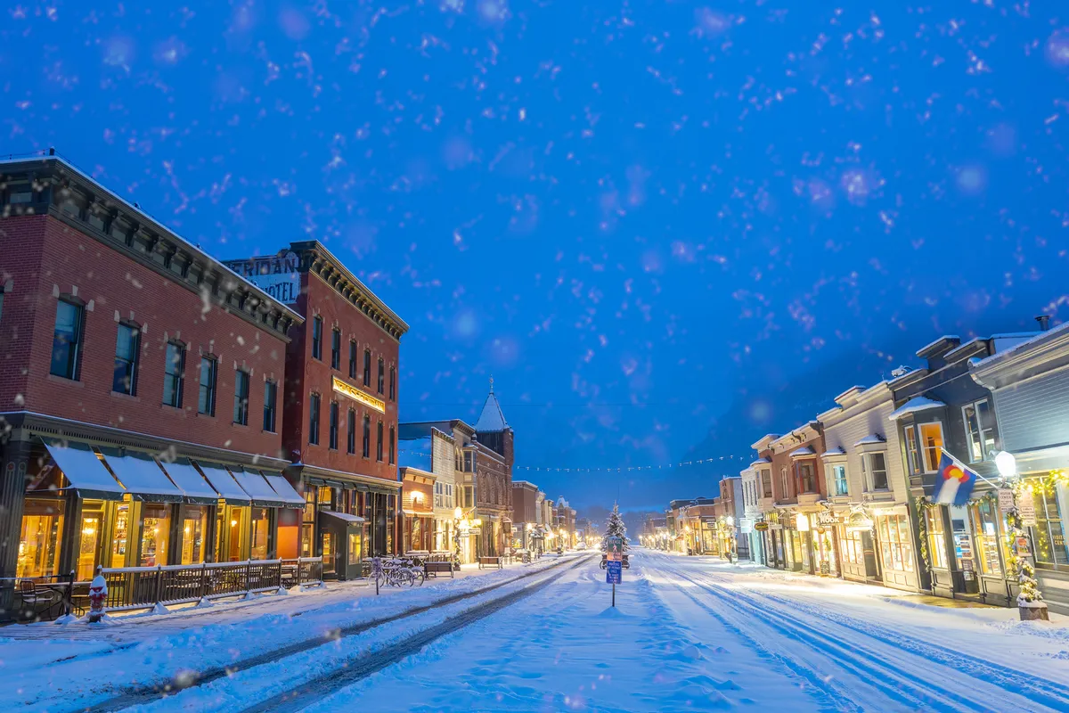 A picturesque, snow-covered street in Telluride lined with illuminated storefronts and adorned with a festive, decorated Christmas tree. People are seen walking and socializing, evoking the charm and vibrancy of the town’s main street during winter.