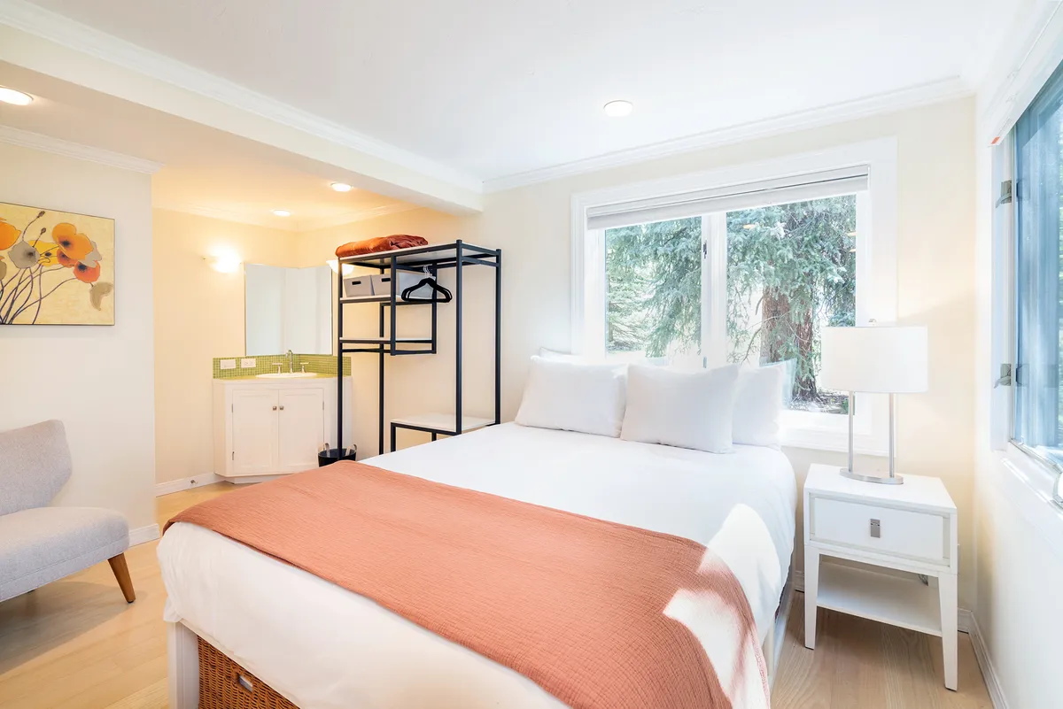 A serene bedroom with a neatly made bed featuring white linens and a coral blanket. Soft natural light streams in through a large window, illuminating the space. Tasteful artwork adds personality, and a modern shelving unit provides convenience and style.
