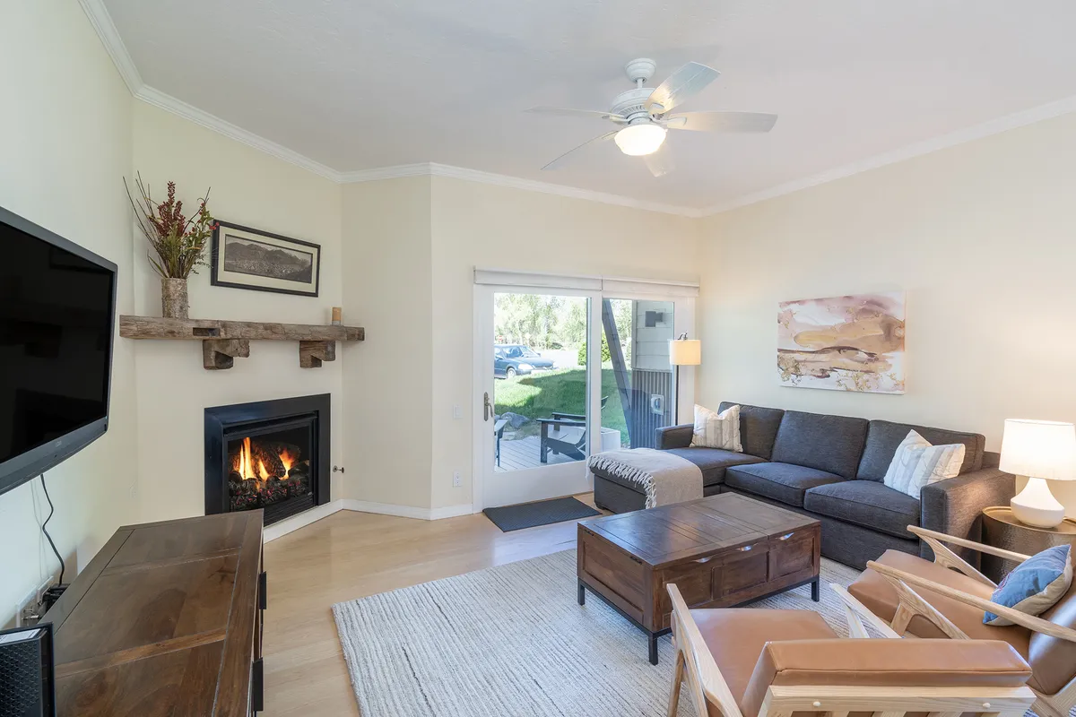 The image showcases a bright, inviting living space centered around a cozy sofa and a glowing fireplace. Sunlight pours in through a large window, illuminating a stylish wooden coffee table and tasteful decorative artwork, creating a comfortable, home-like atmosphere ideal for relaxing or gathering with loved ones.