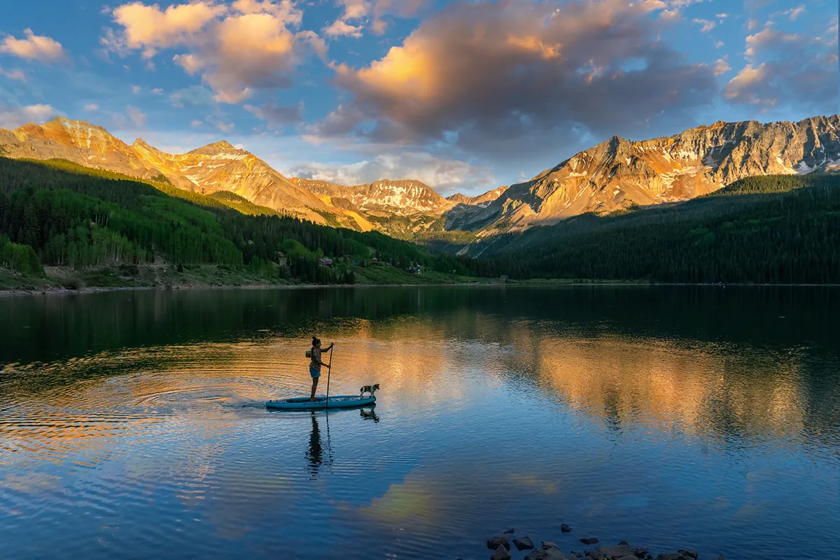 A serene lakeside scene featuring a paddleboarder with their dog, surrounded by mountains and calm water reflecting the landscape. The image evokes relaxation, adventure, and a strong connection to nature.