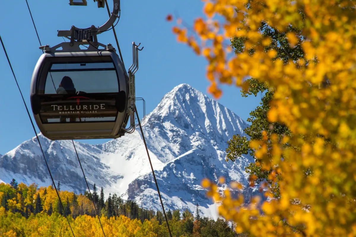 An exterior scene featuring a gondola gliding above snow-capped peaks, with colorful autumn foliage dotting the landscape. The image highlights the adventure and accessibility of Telluride’s outdoors, as well as the stunning year-round beauty of the region.
