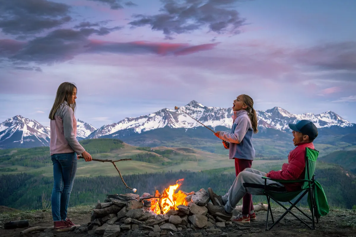 An inviting exterior scene with a campfire as its focal point, surrounded by children roasting marshmallows and an adult relaxing in a chair. The stunning backdrop features a mountain landscape, enhancing the sense of adventure and connection to nature. This image embodies year-round outdoor activity and the family-friendly, communal spirit mentioned in the section.