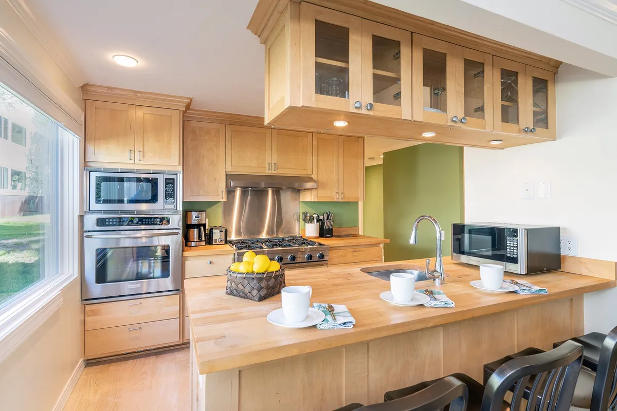 A bright, modern kitchen scene centered around a kitchen island, featuring stainless steel appliances, abundant natural light, and coffee mugs on the counter. The layout invites socializing and casual gatherings, suggesting a space where cooking, brunches, and celebrations happen organically.