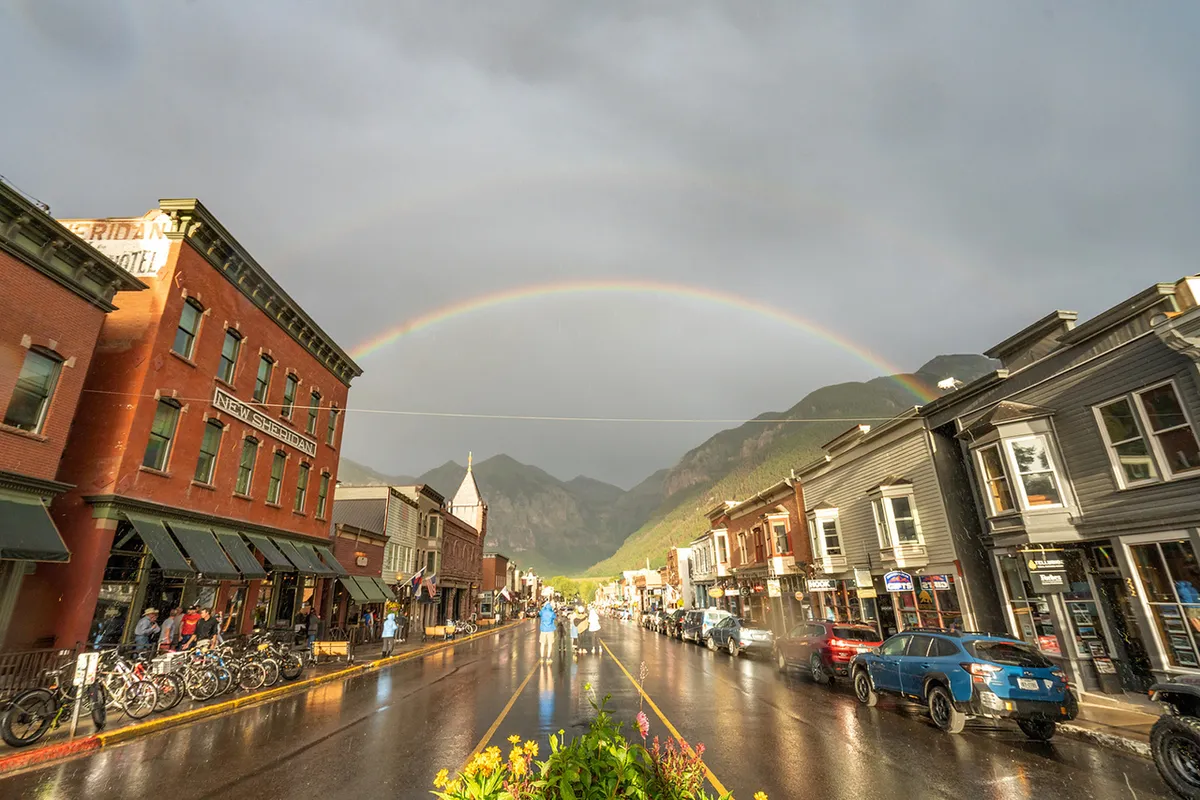 This image depicts Telluride's charming historic downtown, with people exploring local shops, bikes parked along the street, and a rainbow arching overhead. The atmosphere feels vibrant and welcoming, capturing the spirit of adventure and discovery available just outside the doorstep.