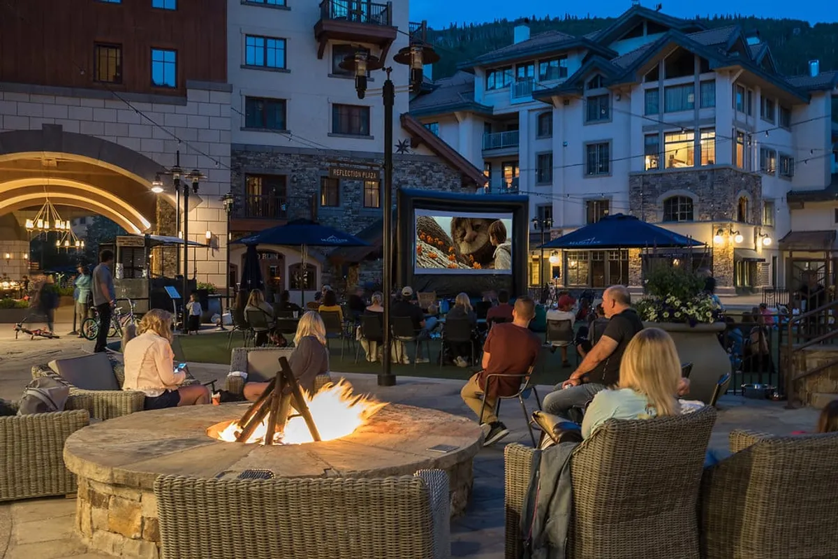 A cozy outdoor fire pit area features a movie screen and comfortable seating, surrounded by modern mountain architecture. The setting offers a communal, relaxing space with scenic views of the mountains in the background, suggesting a perfect place to unwind after a day on the trails.