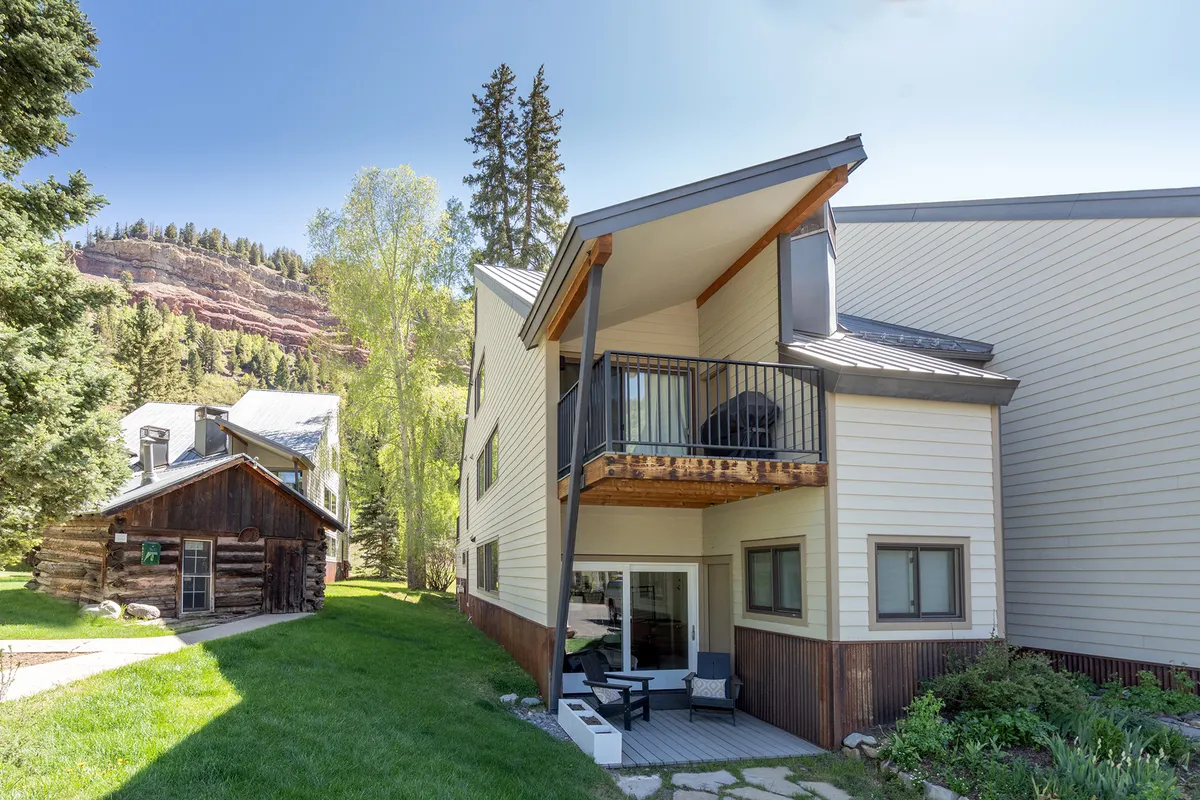 An exterior scene showing a private balcony overlooking a green lawn and surrounded by mature trees, with a view of a nearby wooden cabin. The outdoor space radiates tranquility, offering a secluded spot to enjoy morning coffee, sunsets, or simply the fresh mountain air.