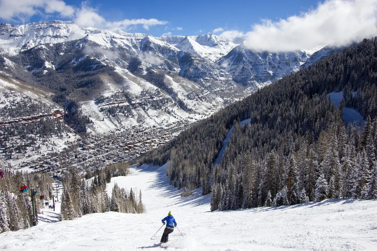 A skier carves down snow-covered slopes against a backdrop of rugged mountains and tall pine trees, with the quaint town visible below. The expansive view captures both the thrill of winter sports and the beauty of the mountain landscape, as well as a sense of proximity between wilderness and home.