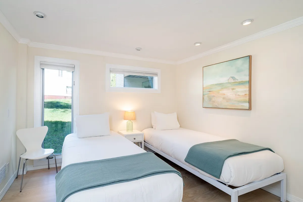 A peaceful bedroom featuring two twin beds separated by a nightstand with a lamp. The room is filled with natural light from a window with a scenic view. Decorative artwork above the beds and simple, functional furnishings create a welcoming environment for families or friends.